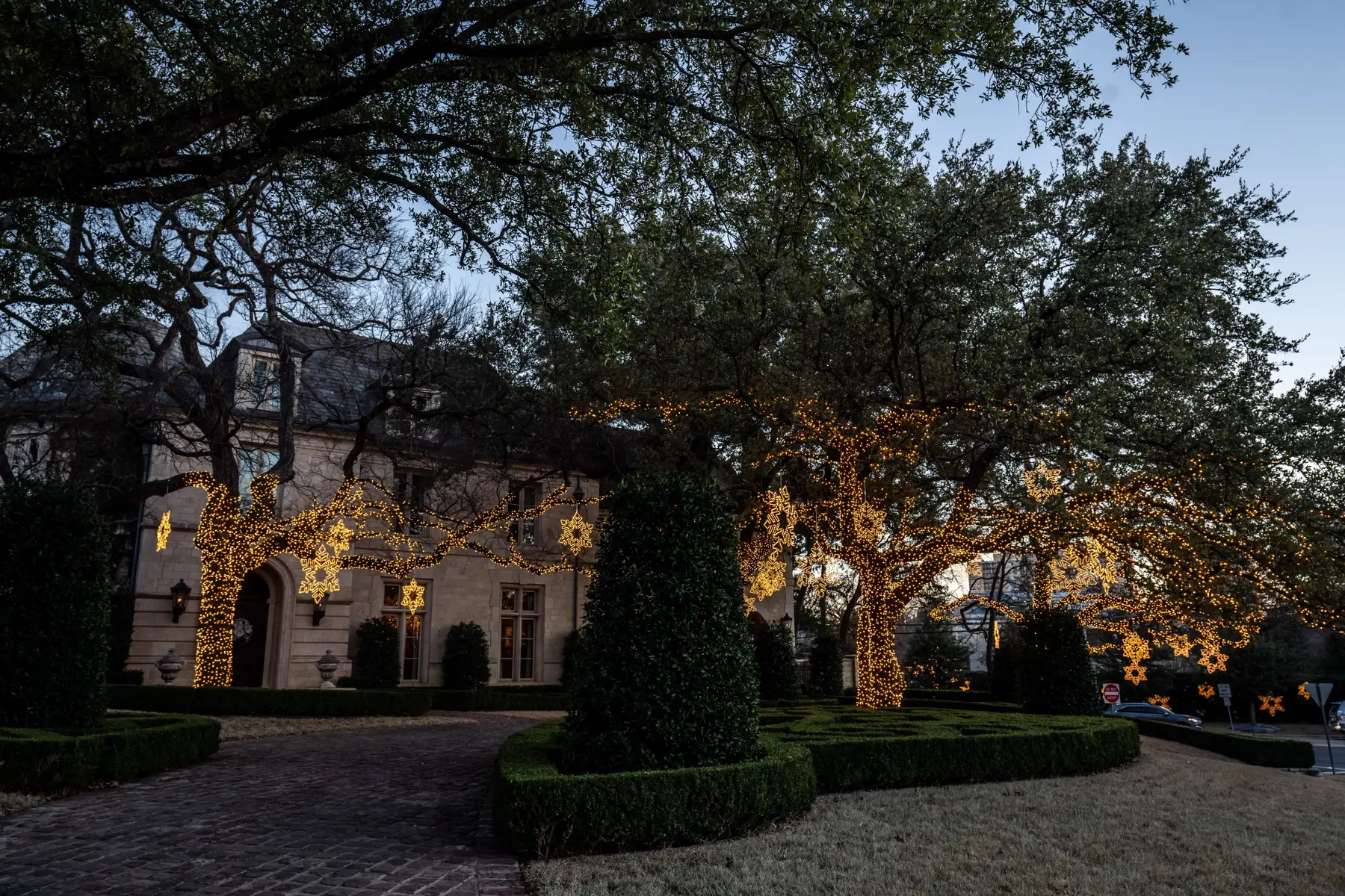 Large home framed by mature trees and landscaping wrapped in warm white holiday lights